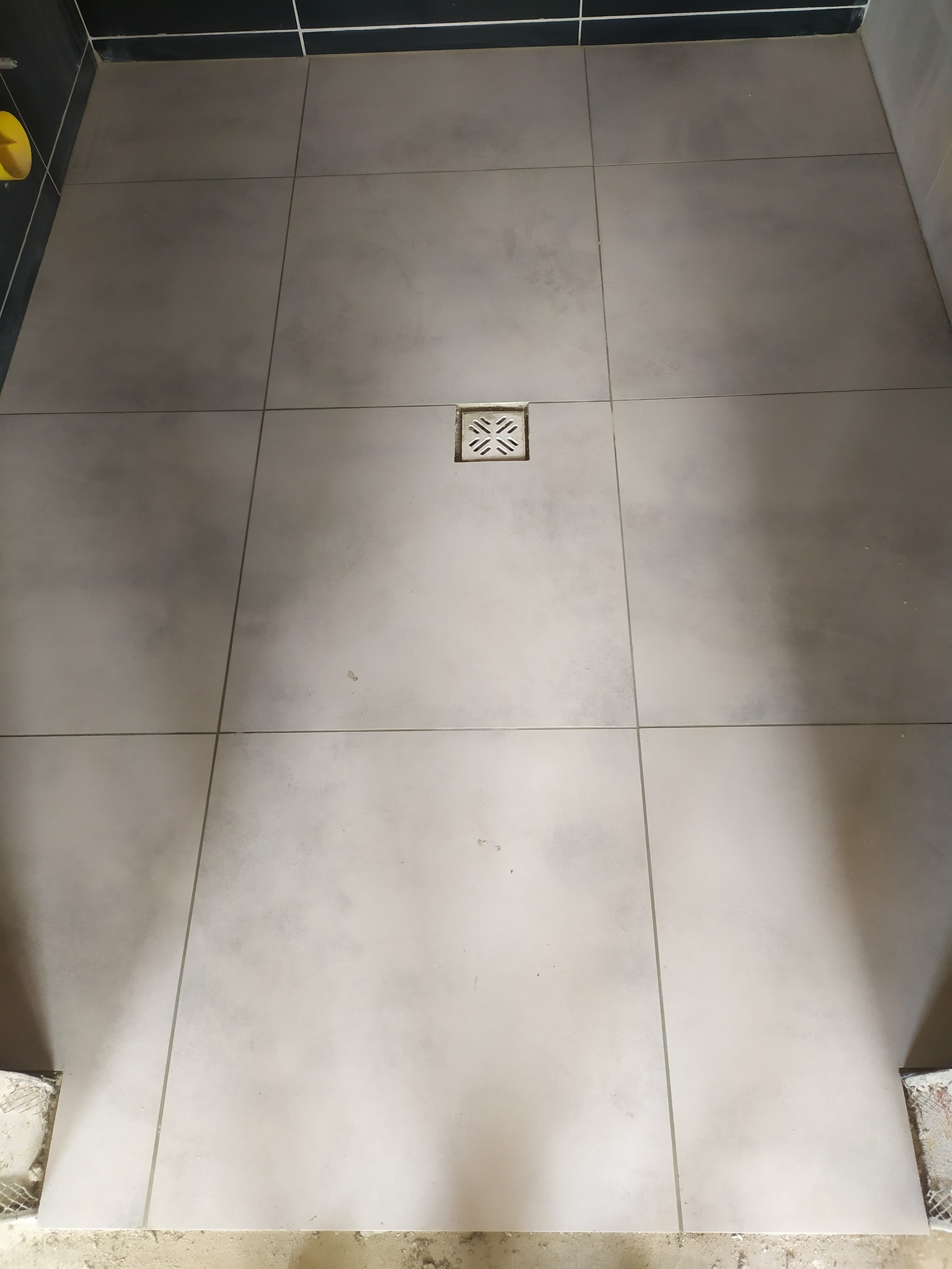 Szare płytki podłogowe w kabinie prysznicowej, z centralnie umieszczonym odpływem liniowym, widoczne niedociągnięcia w wykończeniu przy krawędziach.