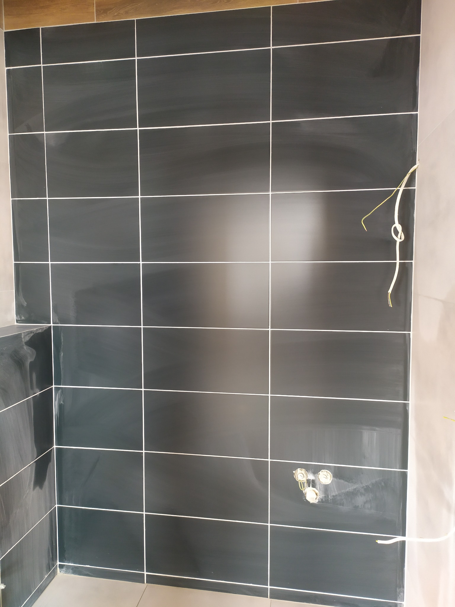 Ściana w łazience wyłożona ciemnymi, błyszczącymi płytkami z jasną fugą, widoczne przygotowane otwory na armaturę i wystające przewody elektryczne.