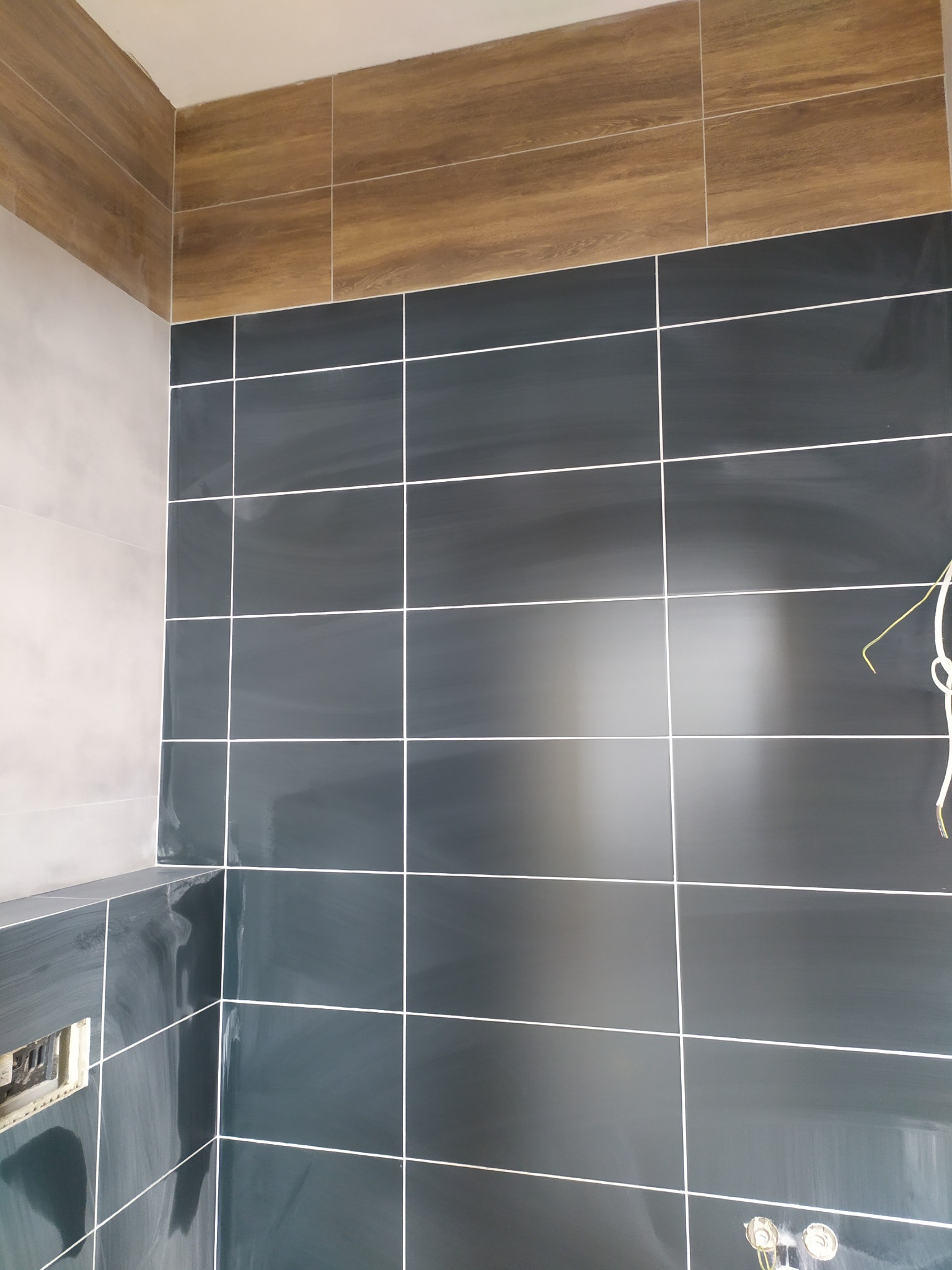 Ściana wyłożona ciemnymi, błyszczącymi płytkami ceramicznymi z białymi fugami, uzupełniona górnym pasem płytek imitujących drewno. Widoczne wystające kable i niedokończone gniazdko elektryczne.