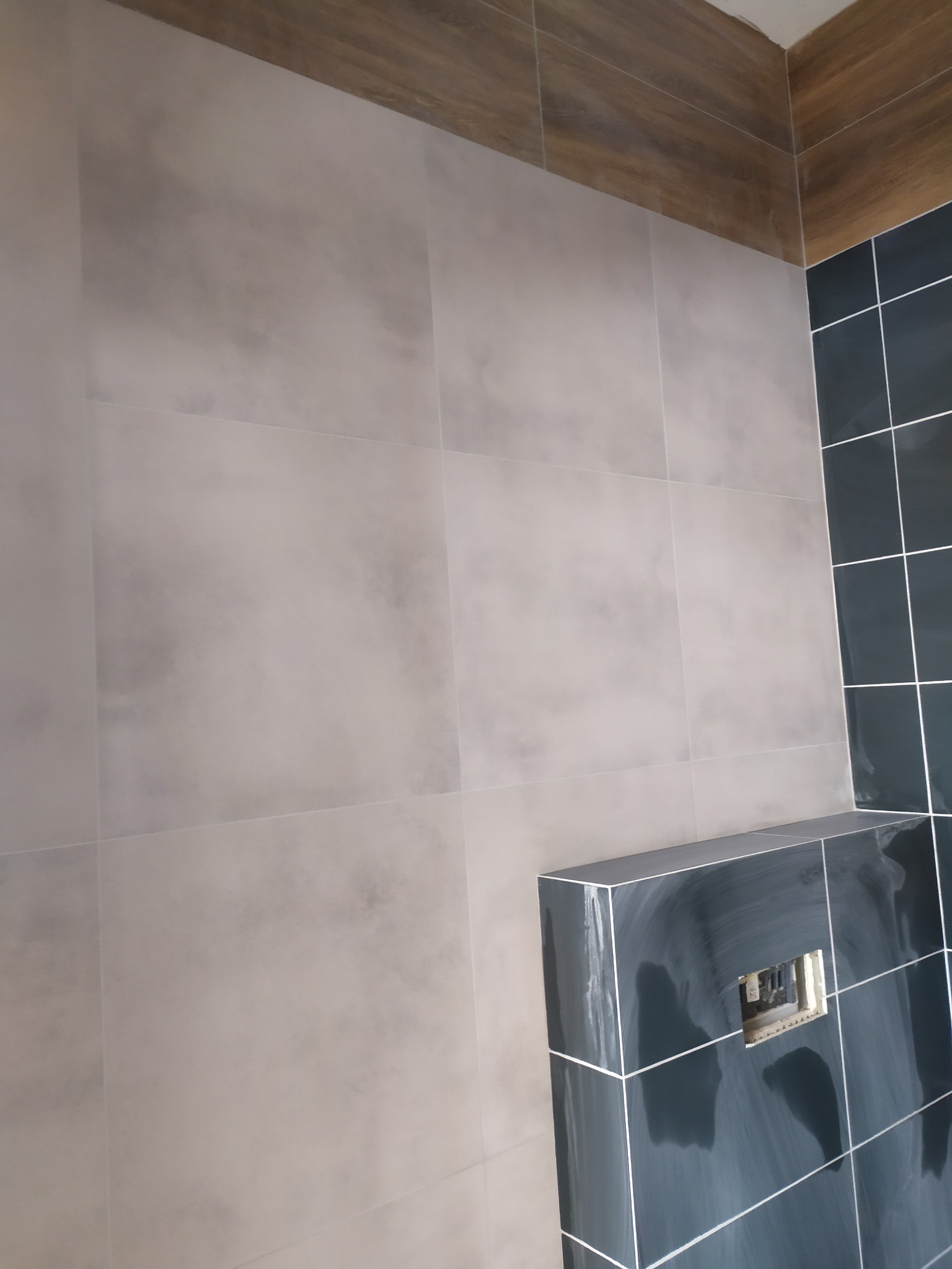 Ściana łazienki wyłożona szarymi płytkami wielkoformatowymi, z fragmentem zabudowy geberitu wykończonej ciemnymi, kwadratowymi płytkami z białą fugą i widocznym otworem na przyłącza.
