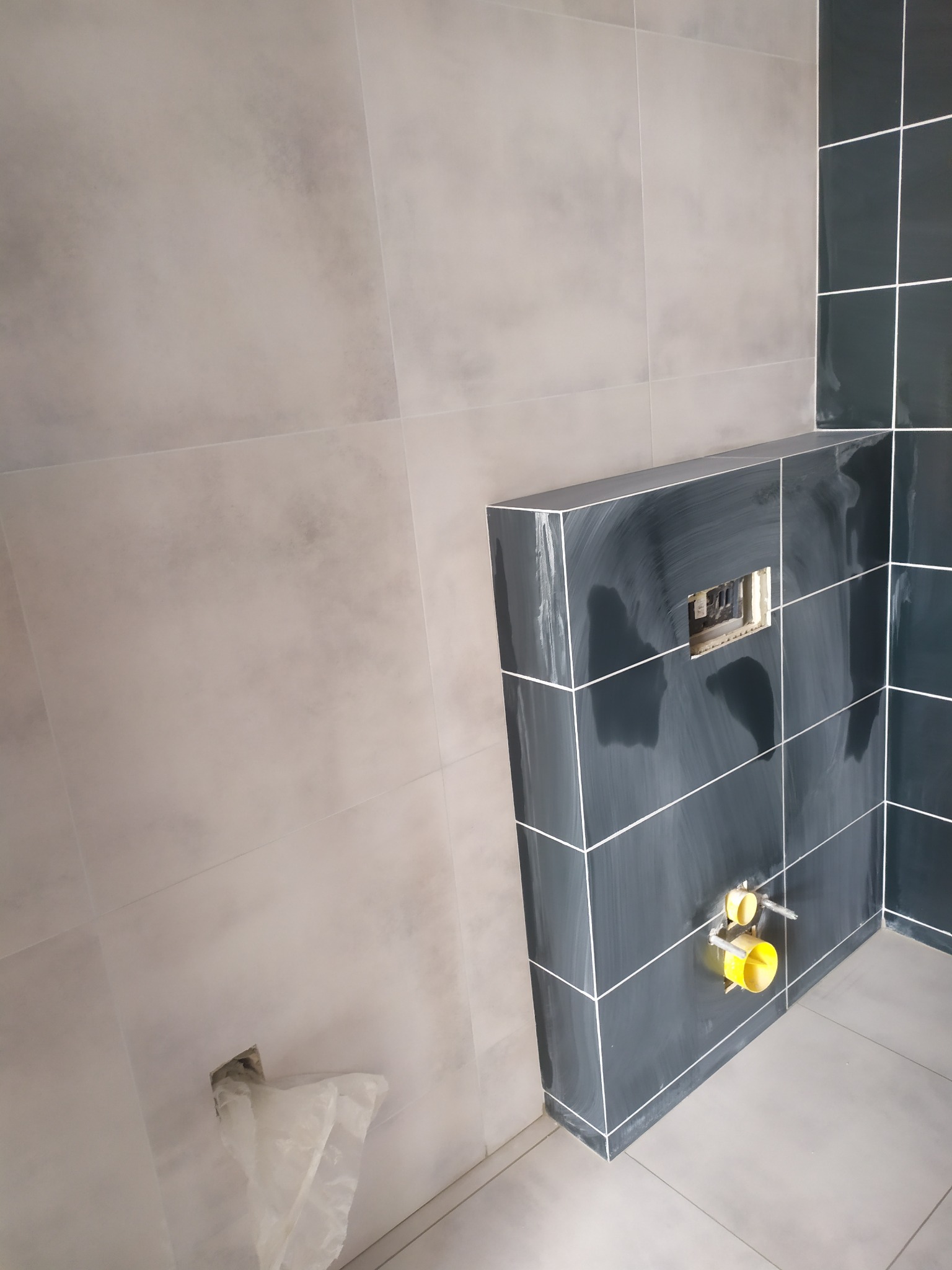 Wnętrze łazienki w trakcie prac wykończeniowych z widocznymi szarymi i grafitowymi płytkami oraz instalacją podtynkową z żółtymi elementami.