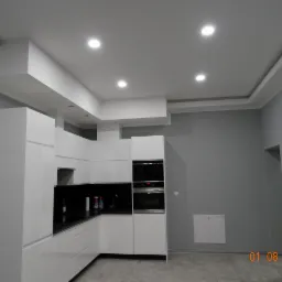 Nowoczesna, biała kuchnia z czarnymi blatami w Bielawie, szare ściany, oświetlenie sufitowe punktowe i dekoracyjny sufit podwieszany.