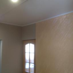 Jacek Altneder Usługi Ogólnobudowlane OTI-BUD - Beżowa ściana z dekoracyjną fakturą, biały sufit z punktowym oświetleniem i drewniane drzwi z szybami w odcieniach beżu.