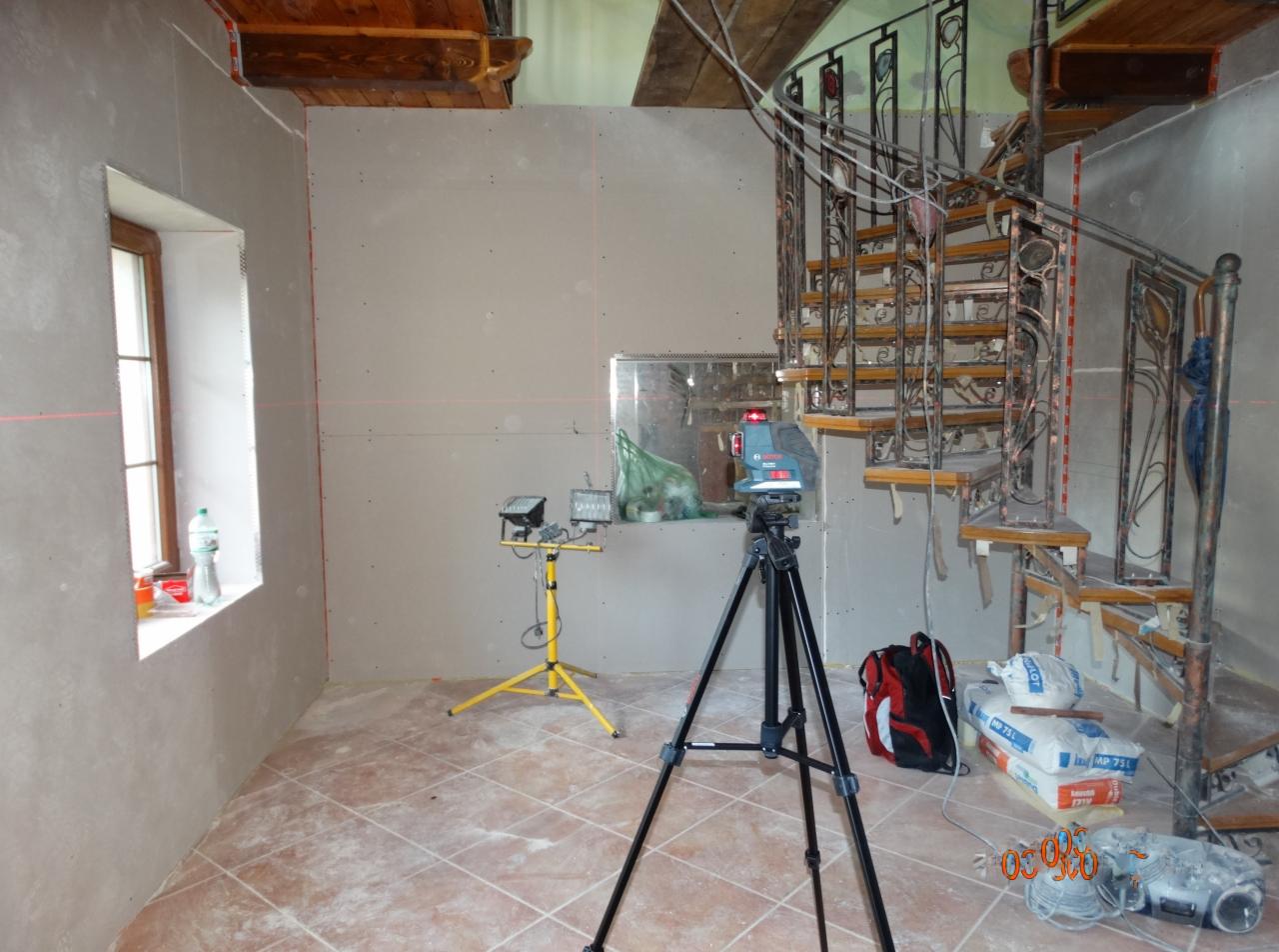 Wnętrze w trakcie prac wykończeniowych: ściany pokryte płytami gipsowo-kartonowymi, poziomica laserowa na statywie, reflektory na statywie, spiralne schody z metaloplastyki, worki z zaprawą gipsową...