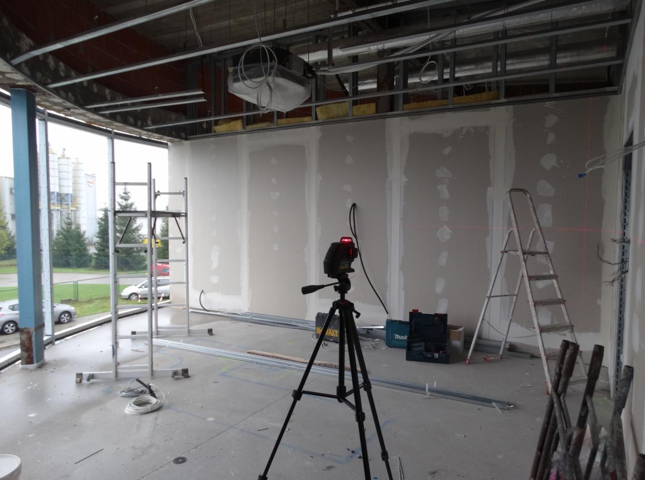 Wnętrze w trakcie prac wykończeniowych: ściany z płyt gipsowo-kartonowych, laser krzyżowy na statywie wyznacza poziom, widoczne drabiny, profile metalowe i narzędzia.