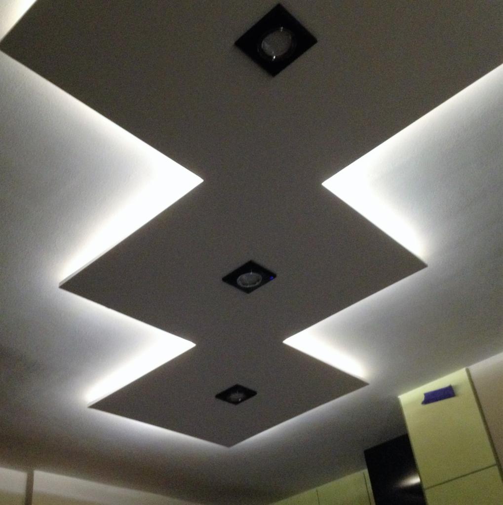 Kaskadowy sufit podwieszany z oświetleniem LED tworzącym geometryczny wzór, trzy kwadratowe lampy punktowe w czarnych oprawach, widok z dołu.