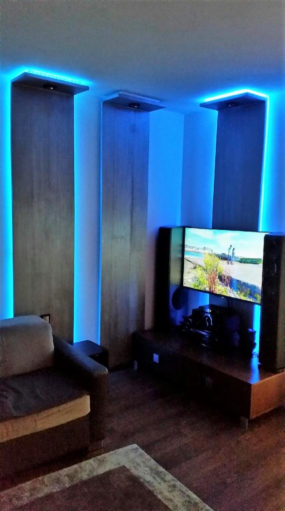 Nowoczesny salon z podświetlanymi panelami ściennymi i sufitowymi, oświetlenie LED w kolorze niebieskim, telewizor z włączonym ekranem, drewniana podłoga i szary narożnik.