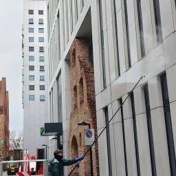 AquaUp - Pracownik w zielonej kurtce czyści elewację budynku za pomocą lancy teleskopowej. W tle nowoczesna architektura i fragment zabytkowej, ceglanej budowli.