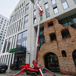 AquaUp - Czerwony podnośnik koszowy na gąsienicach przed budynkiem z ceglaną fasadą i nowoczesną częścią. Widoczny kosz na wysokości okien.