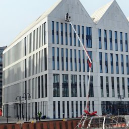 AquaUp - Pracownik w koszu podnośnika czyści okna nowoczesnego biurowca z charakterystycznym szczytem dachu, widok z nabrzeża z łodzią i osobą w kamizelce odblaskowej.