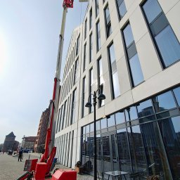 AquaUp - Czerwony podnośnik koszowy przy nowoczesnym, białym budynku z licznymi oknami. Widoczne ślady mycia na elewacji i oknach. W tle zabytkowa architektura miejska.