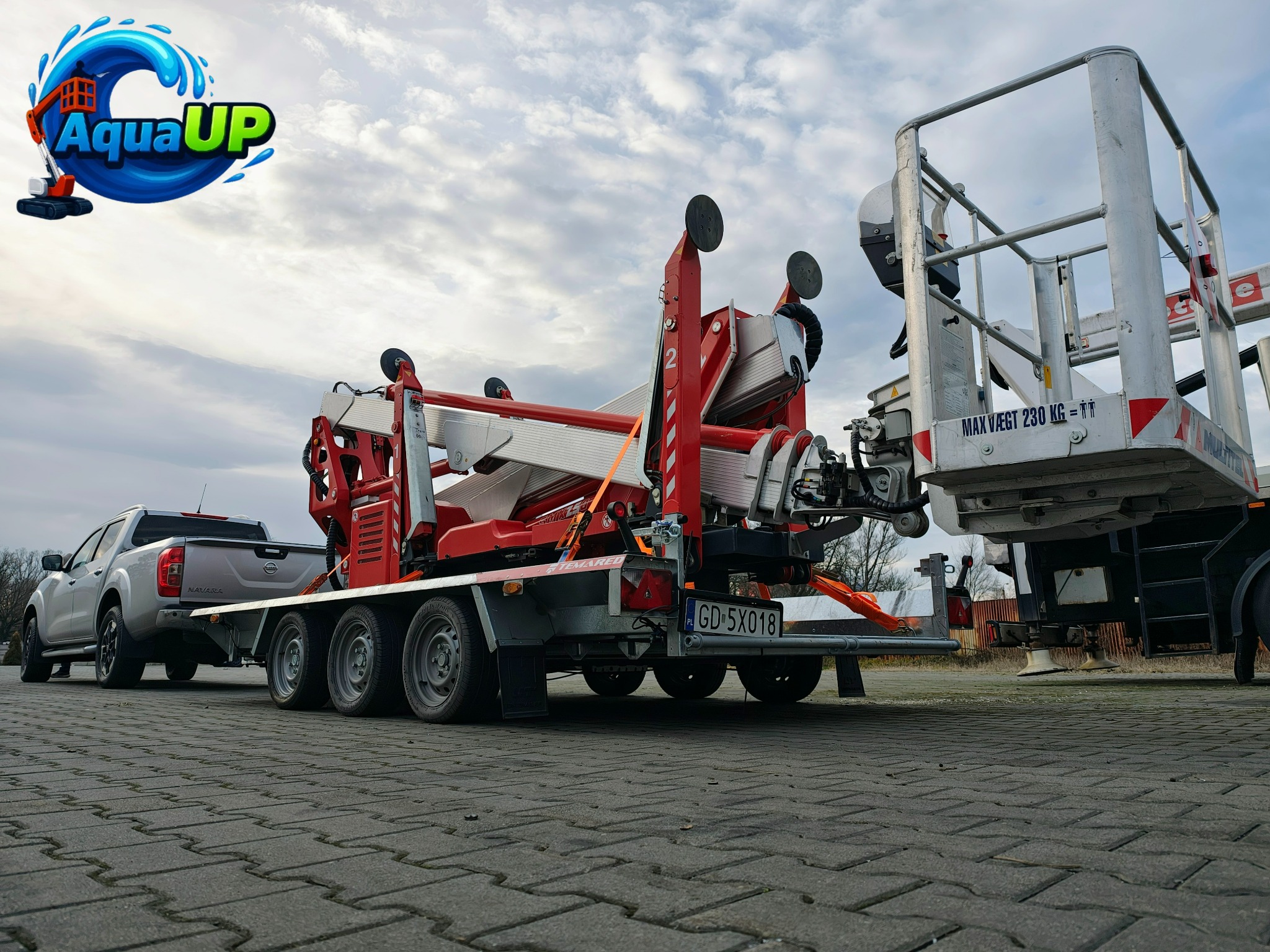 Podnośnik koszowy na przyczepie, z wysięgnikiem w kolorach czerwieni i bieli, zaparkowany na brukowanej powierzchni, z szarym pickupem w tle.