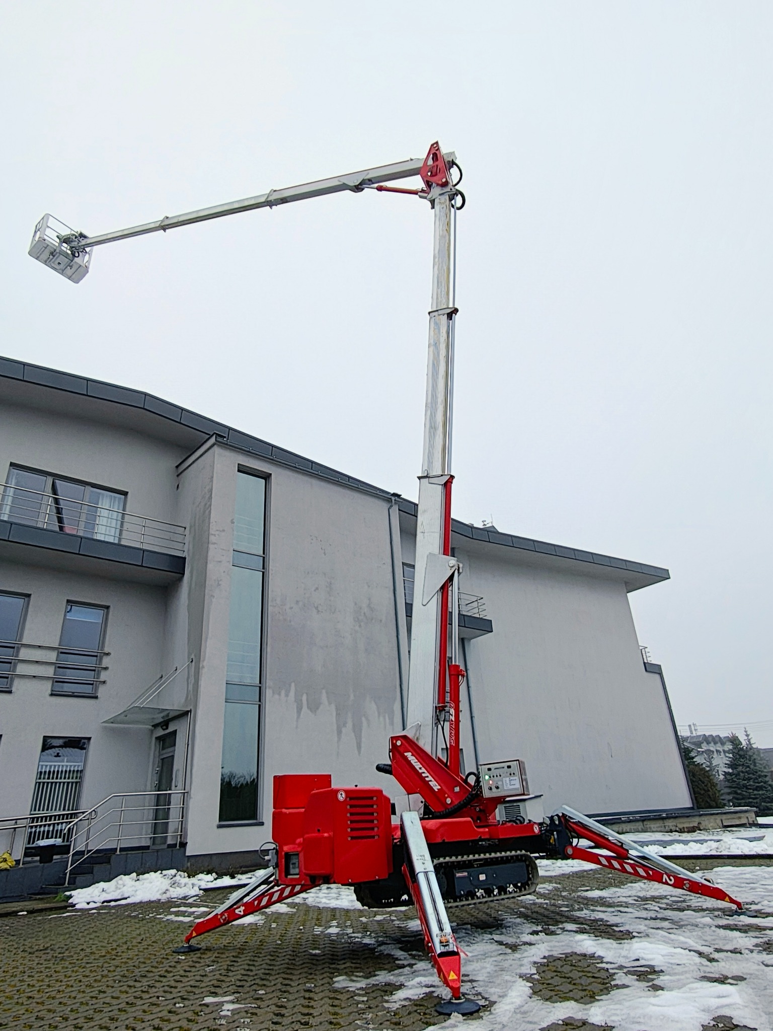 Czerwony podnośnik koszowy na gąsienicach, z wysięgnikiem i koszem, ustawiony na kostce brukowej przed budynkiem w pochmurny dzień. Widoczne podpory stabilizujące.
