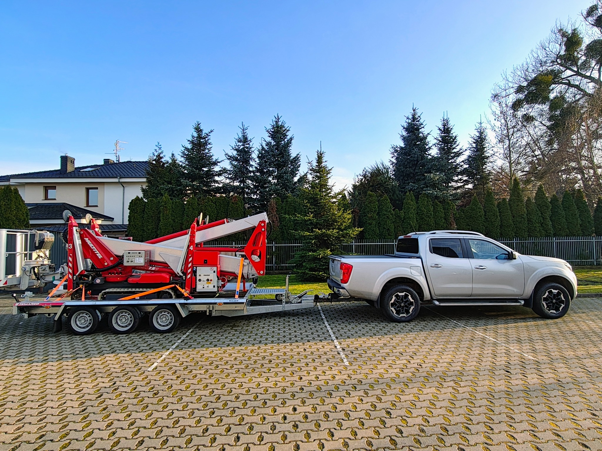 Podnośnik koszowy na przyczepie, ciągnięty przez srebrnego pickupa na parkingu z kostki. W tle budynek i drzewa pod błękitnym niebem.