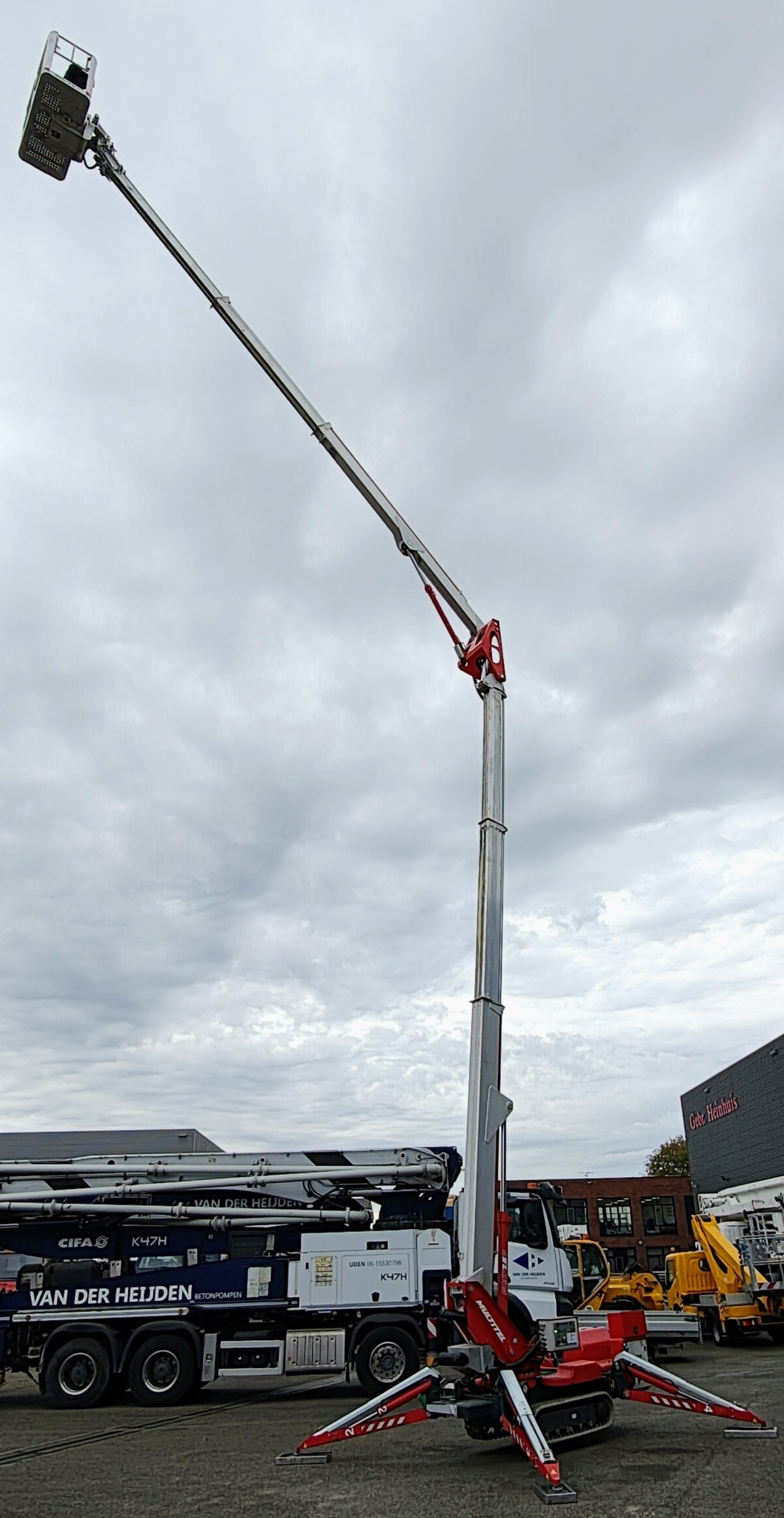 Podnośnik koszowy z wysięgnikiem teleskopowym na tle pochmurnego nieba, zaparkowany obok betonomieszarki i innych pojazdów budowlanych.