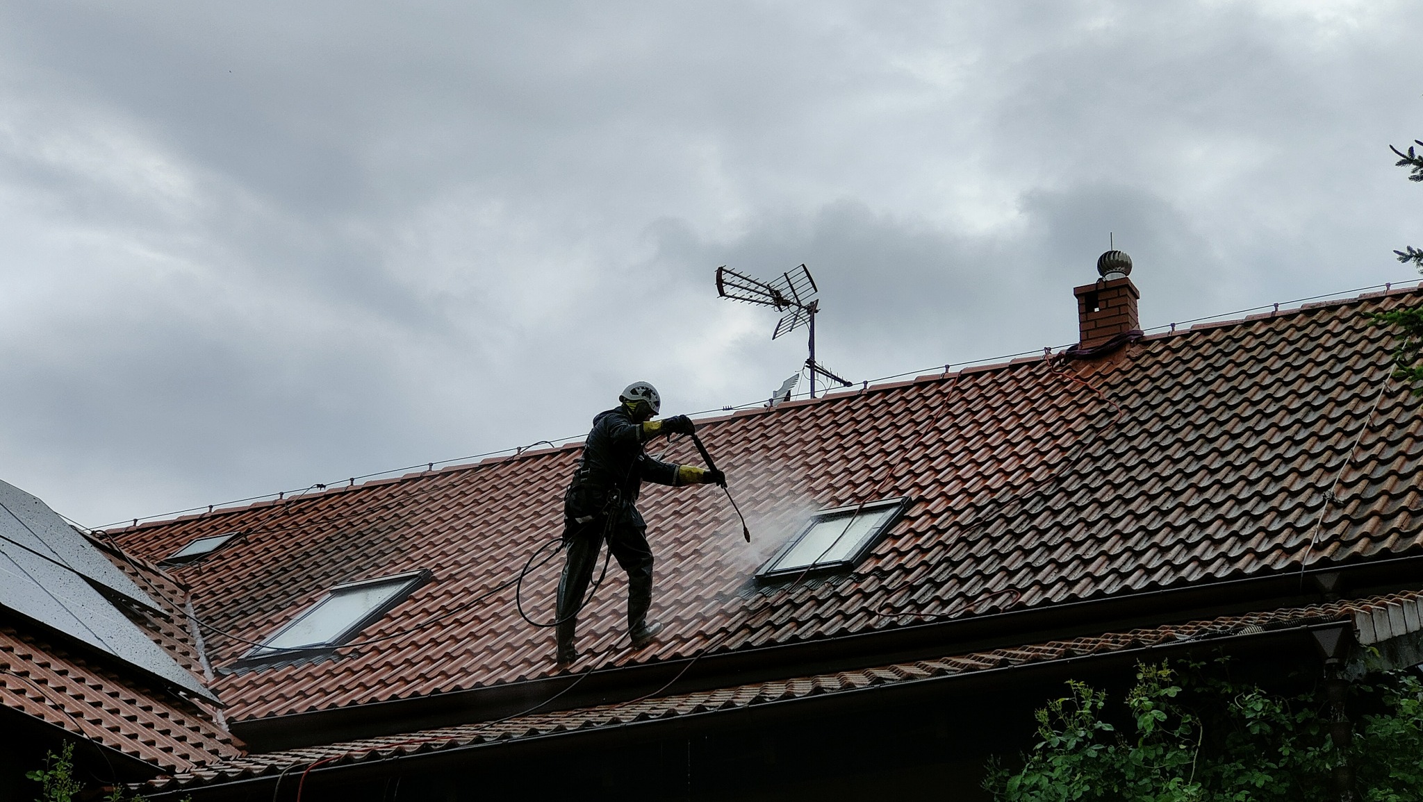 Pracownik w kombinezonie i kasku czyści dachówkę z mchu i zabrudzeń za pomocą myjki ciśnieniowej. Widoczne okna dachowe, antena i komin na tle pochmurnego nieba.