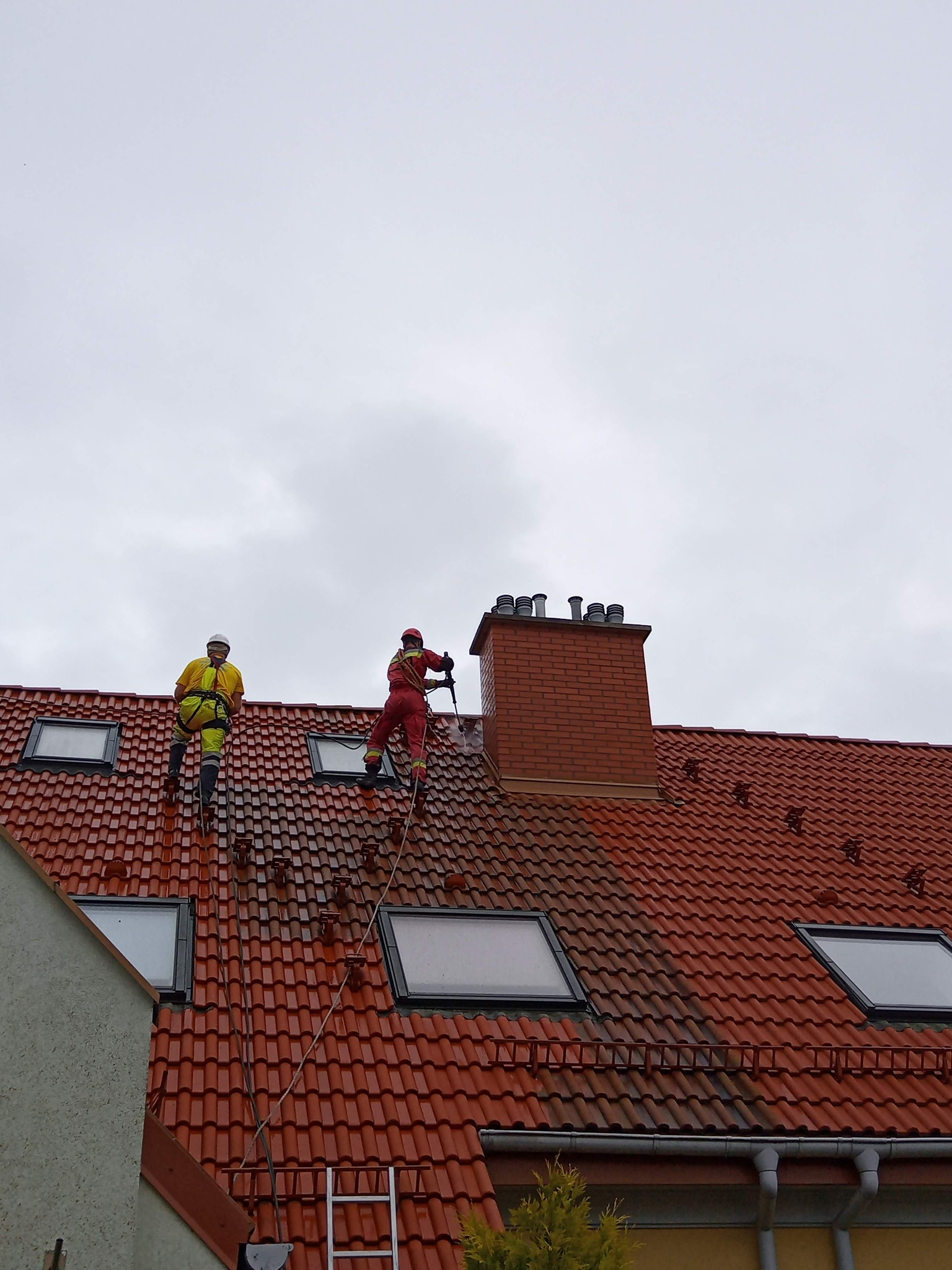 Dwóch pracowników w kombinezonach czyści czerwoną dachówkę z użyciem myjki ciśnieniowej. Widoczny komin i okna dachowe. Część dachu jest już umyta, ukazując różnicę.