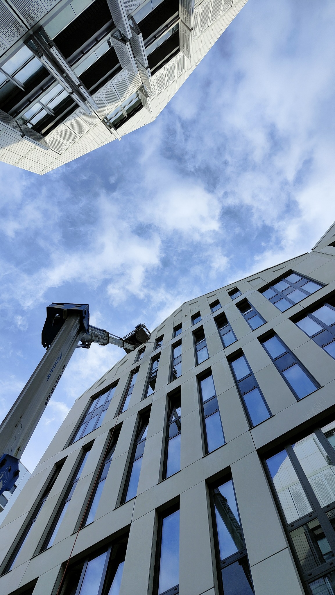 Elewacja budynku z podnośnikiem koszowym na tle błękitnego nieba. Widok z dołu, podkreślający wysokość i skalę prac. Nowoczesna architektura.