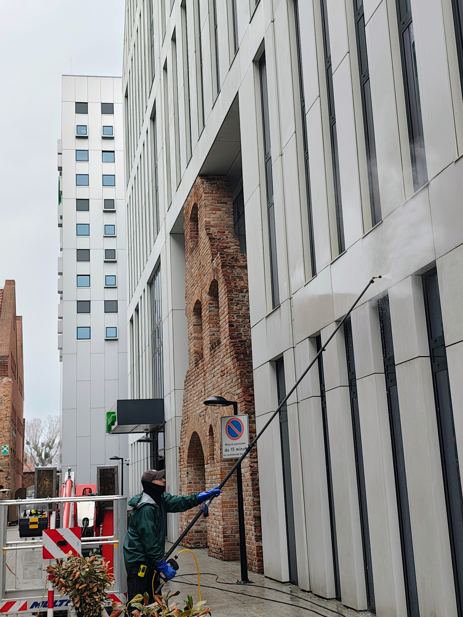 Pracownik w zielonej kurtce czyści elewację budynku za pomocą lancy teleskopowej. W tle nowoczesna architektura i fragment zabytkowej, ceglanej budowli.