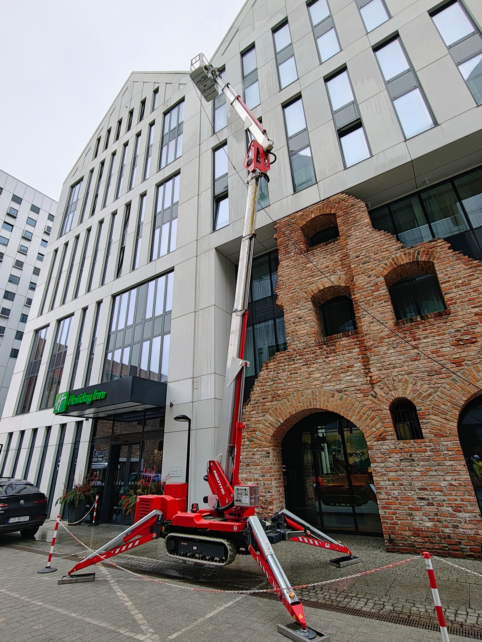 Czerwony podnośnik koszowy na gąsienicach przed budynkiem z ceglaną fasadą i nowoczesną częścią. Widoczny kosz na wysokości okien.