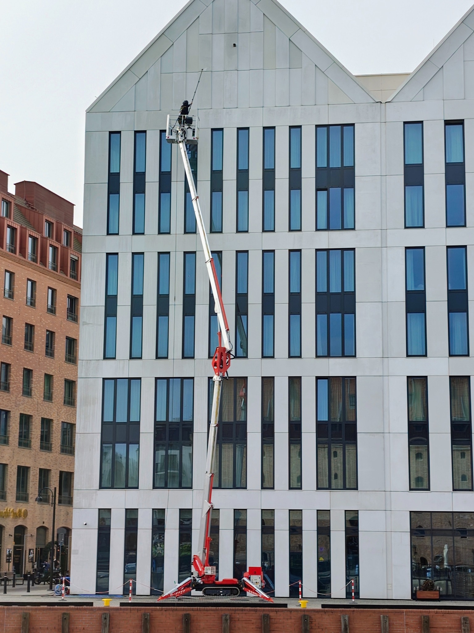 Pracownik w koszu podnośnika czyści elewację nowoczesnego budynku z licznymi, wąskimi oknami. W tle widoczny budynek z cegły i fragment nabrzeża.
