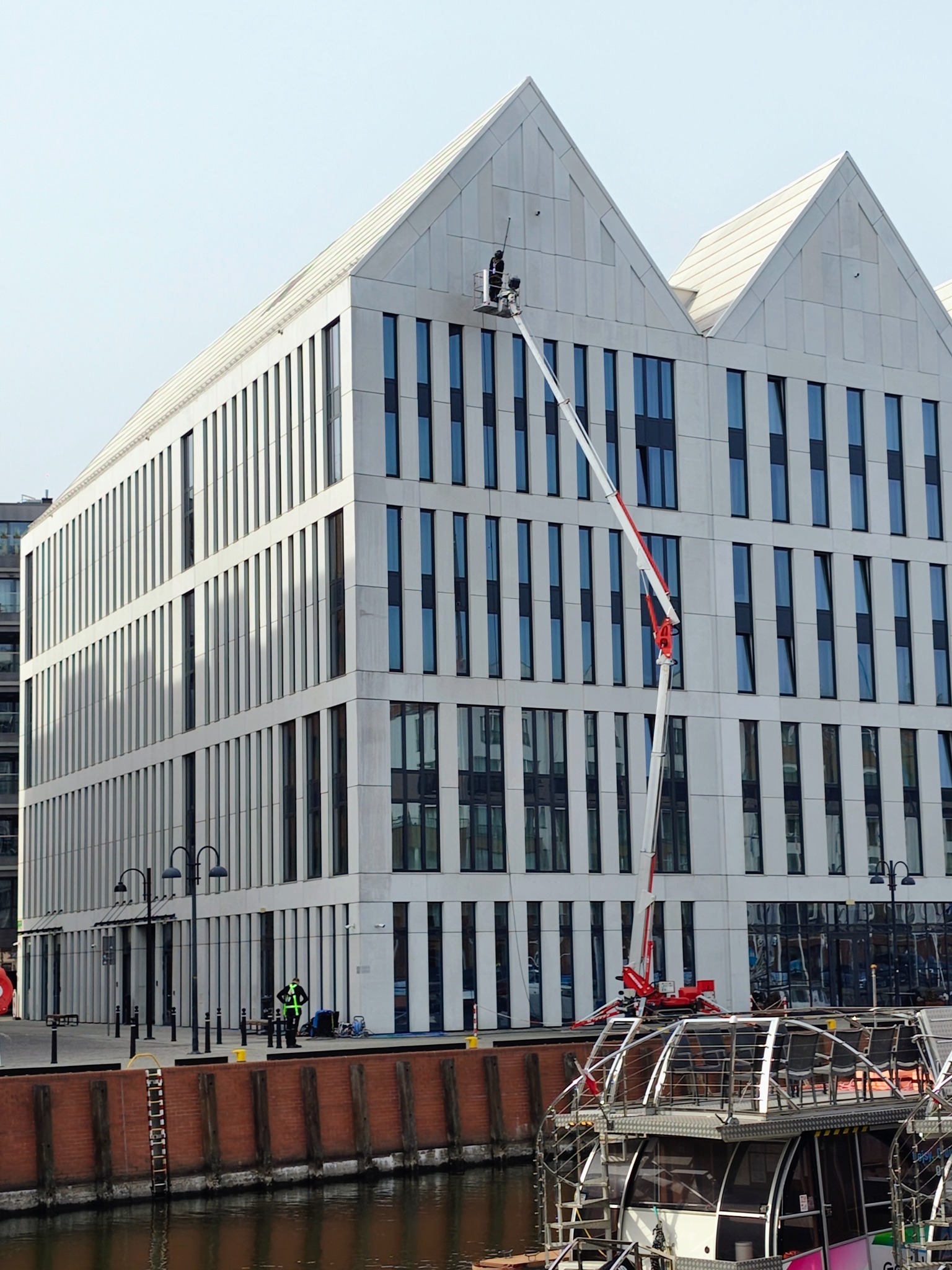 Pracownik w koszu podnośnika czyści okna nowoczesnego biurowca z charakterystycznym szczytem dachu, widok z nabrzeża z łodzią i osobą w kamizelce odblaskowej.