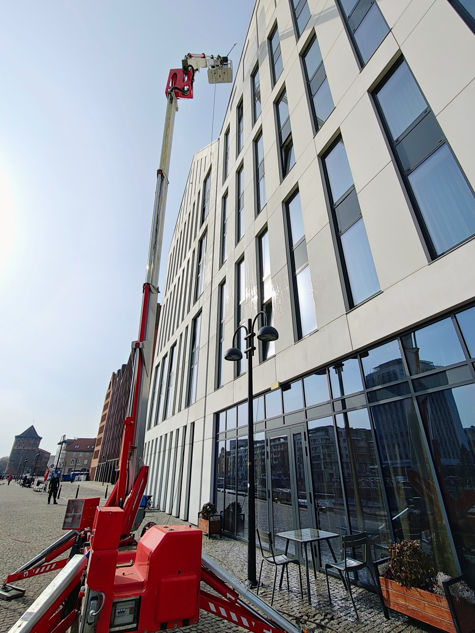 Czerwony podnośnik koszowy przy nowoczesnym, białym budynku z licznymi oknami. Widoczne ślady mycia na elewacji i oknach. W tle zabytkowa architektura miejska.