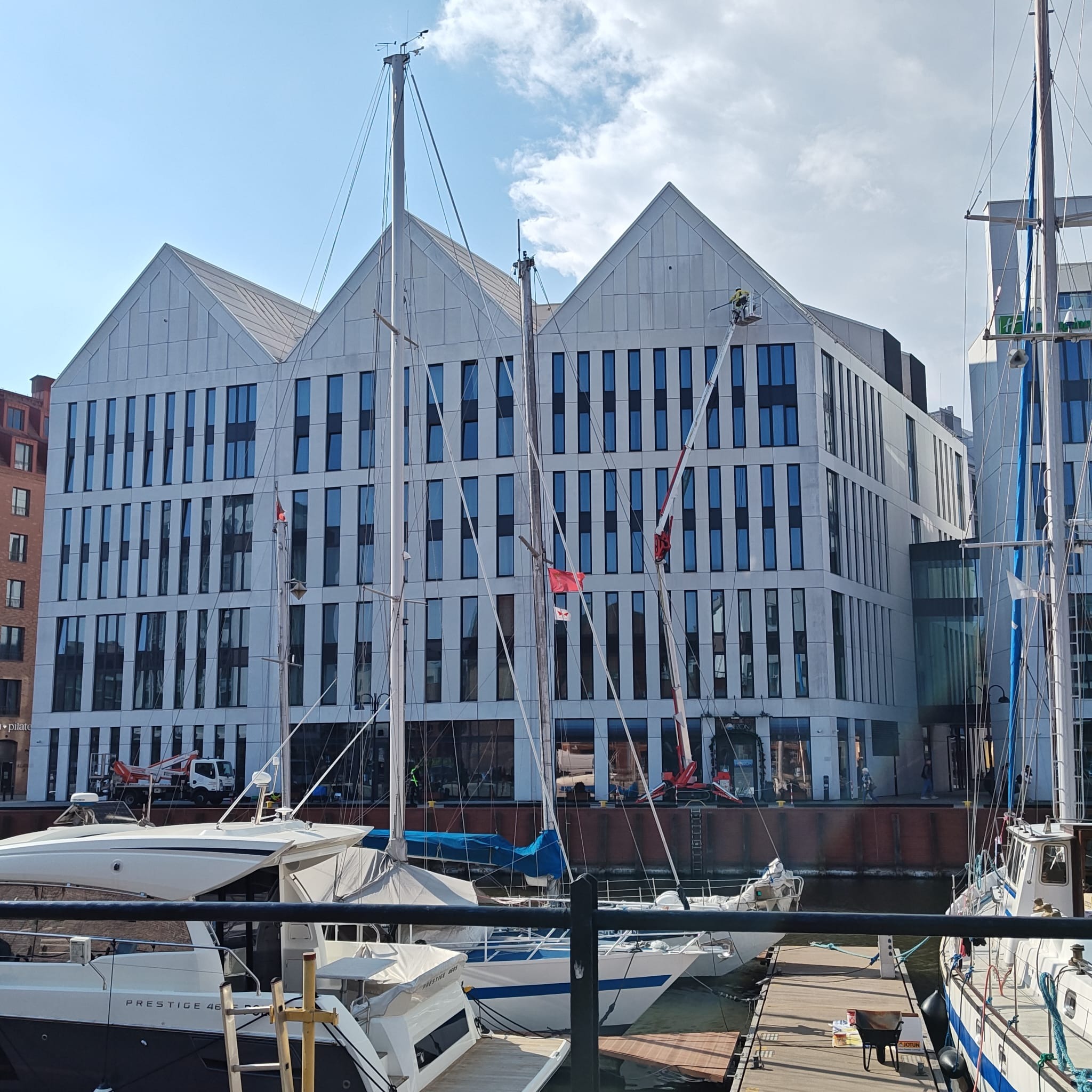 Mycie okien na wysokości w nowoczesnym budynku z podnośnika koszowego, na pierwszym planie jachty w porcie, słoneczny dzień.