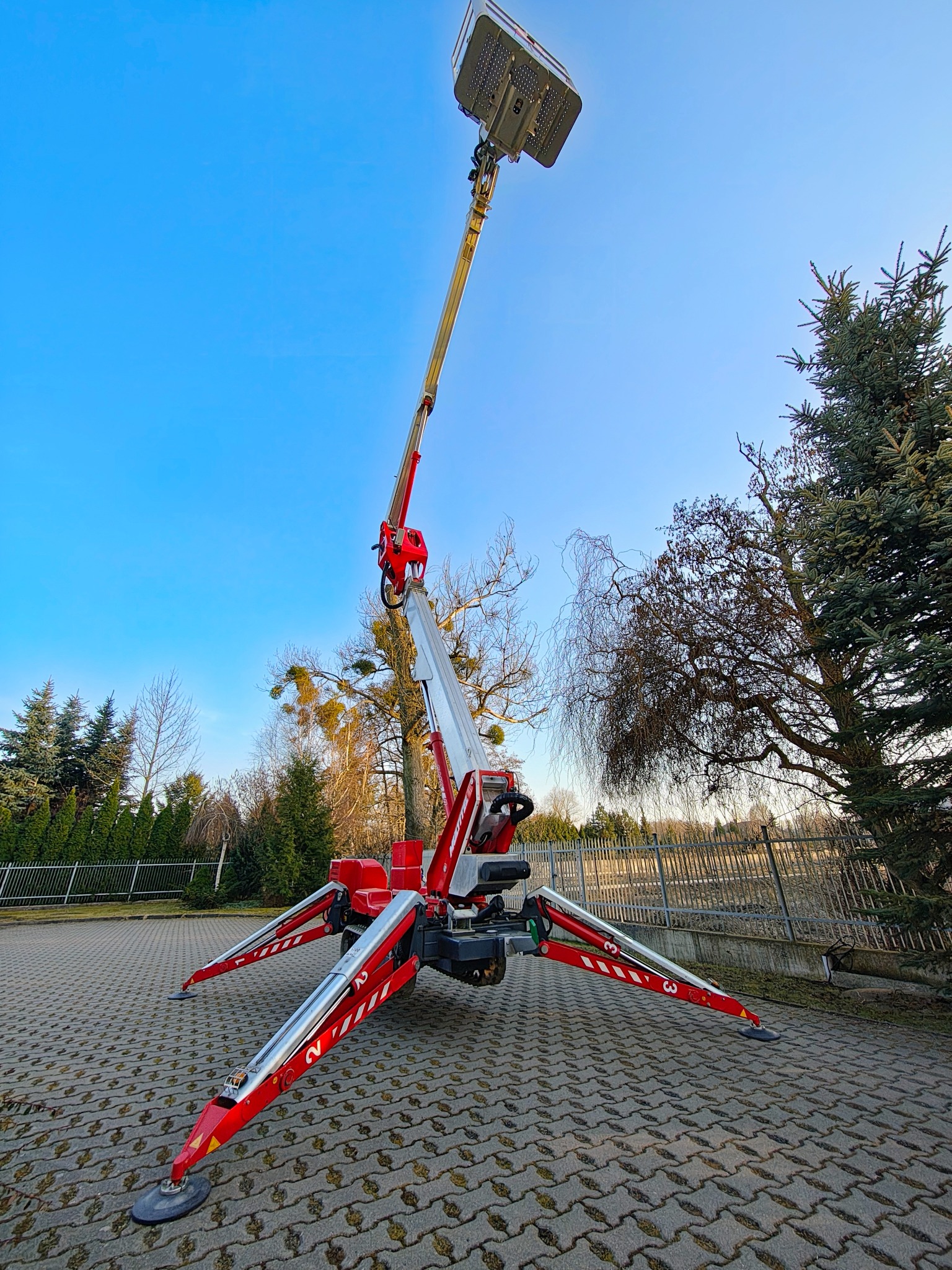 Czerwono-srebrny podnośnik koszowy z wysięgnikiem na tle błękitnego nieba i drzew. Widok z dołu, ukazujący konstrukcję i stabilizatory z numerami.