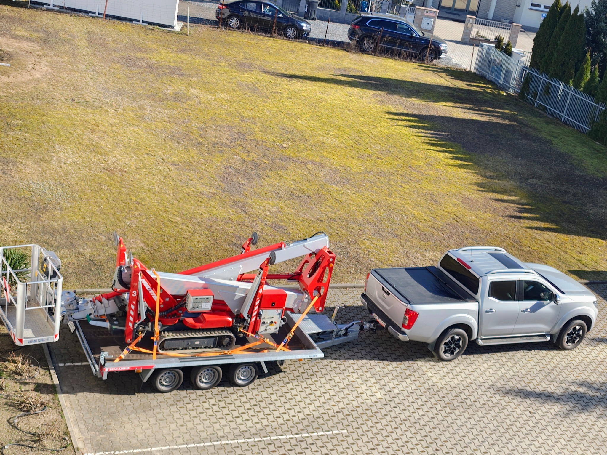 Ciężarówka z platformą na przyczepie, na której znajduje się czerwony podnośnik koszowy gąsienicowy. Widok z góry na plac z kostki brukowej i trawnik w tle.