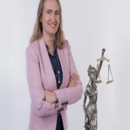 Portret uśmiechniętej kobiety w różowym żakiecie i granatowych spodniach, stojącej z założonymi rękami obok figury Temidy na białym tle.