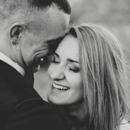 Czarno-białe zbliżenie uśmiechniętej kobiety z zamkniętymi oczami, wtulonej w mężczyznę w garniturze, delikatne oświetlenie, logo w prawym górnym rogu.