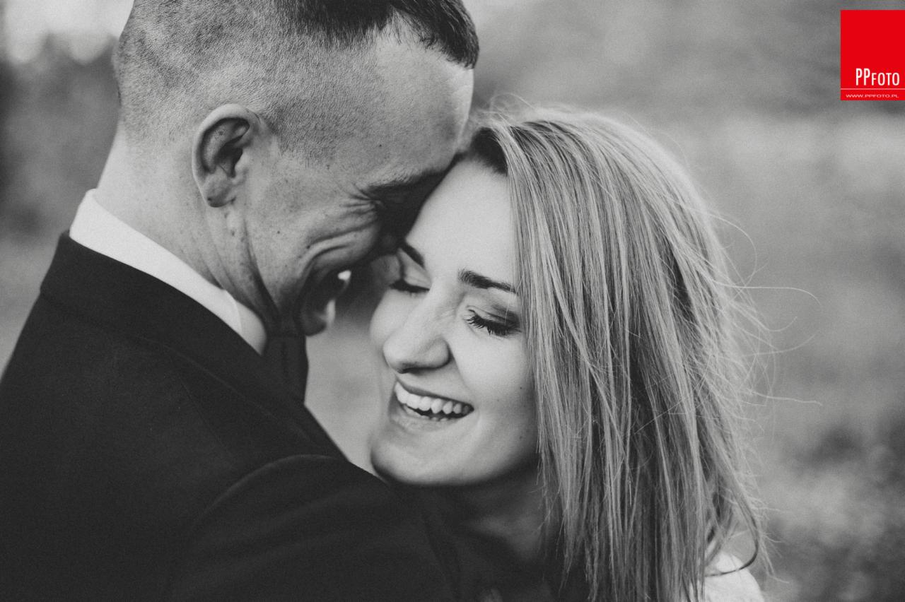 Czarno-białe zbliżenie uśmiechniętej kobiety z zamkniętymi oczami, wtulonej w mężczyznę w garniturze, delikatne oświetlenie, logo w prawym górnym rogu.