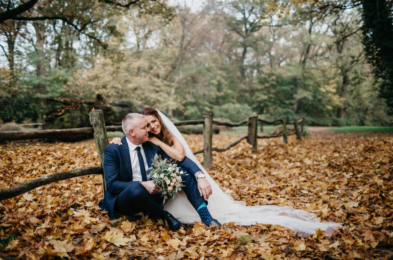 Para młoda siedzi na ziemi pokrytej jesiennymi liśćmi, opierając się o drewniany płot. Panna młoda w białej sukni i welonie, z bukietem w ręku, przytula się do pana młodego w granatowym garniturze...