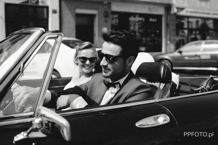 Czarno-białe zdjęcie uśmiechniętej pary młodej w kabriolecie, mężczyzna w garniturze i muszce prowadzi auto, kobieta w sukni ślubnej siedzi obok, oboje w okularach przeciwsłonecznych.