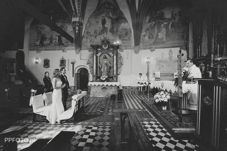Czarno-białe zdjęcie pary młodej stojącej przed ołtarzem w kościele z freskami na ścianach i posadzką w szachownicę, w tle ksiądz i ministranci.