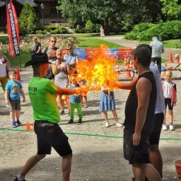 Mężczyzna w kapeluszu i zielonej koszulce z napisem 'Animator' przekazuje płomień ognia drugiemu mężczyźnie, otoczeni przez grupę dzieci i dorosłych w parku.