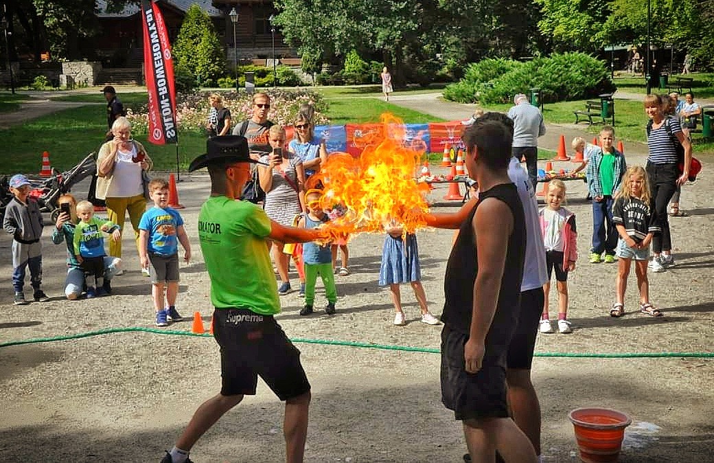 Mężczyzna w kapeluszu i zielonej koszulce z napisem 'Animator' przekazuje płomień ognia drugiemu mężczyźnie, otoczeni przez grupę dzieci i dorosłych w parku.
