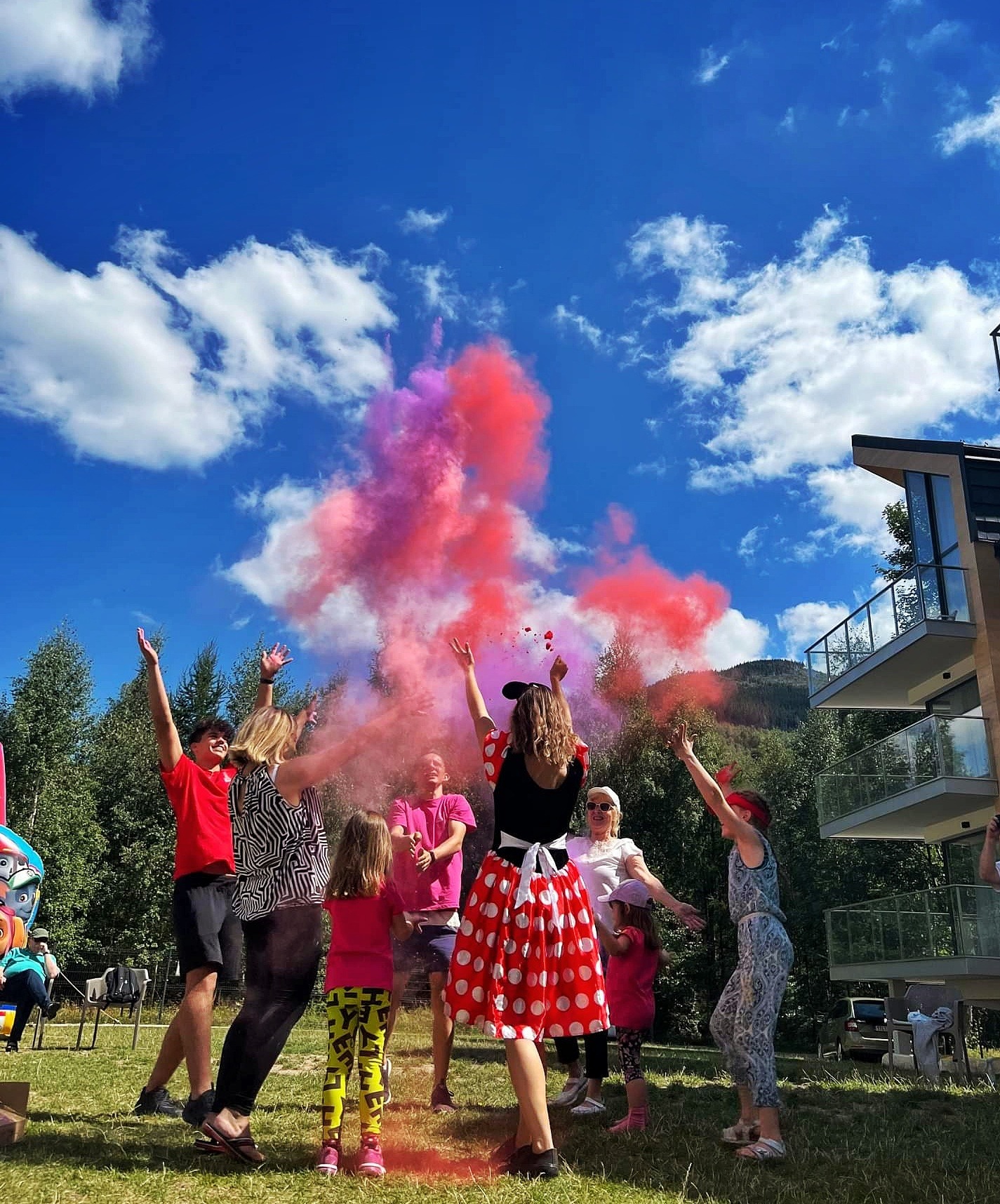 Animator w stroju Myszki Minnie i grupa dzieci oraz dorosłych rzucają kolorowym proszkiem holi na tle błękitnego nieba z białymi chmurami i nowoczesnego budynku.