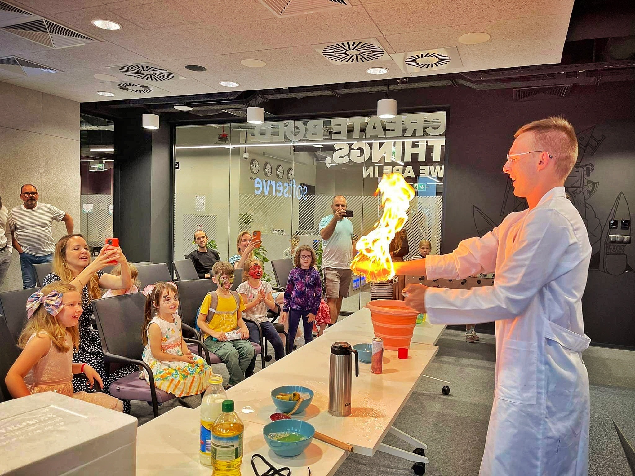 Naukowiec w białym fartuchu prezentuje eksperyment z ogniem dla dzieci z pomalowanymi twarzami, siedzących na krzesłach w nowoczesnym pomieszczeniu.