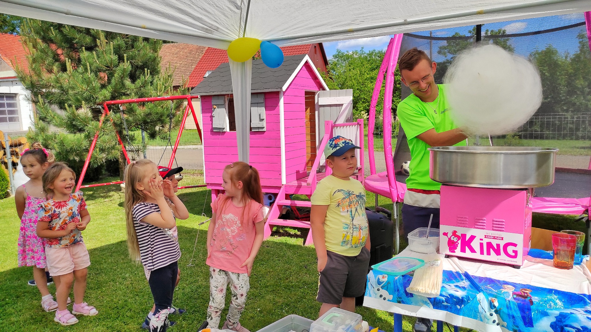 Mężczyzna w okularach i jaskrawozielonej koszulce robi watę cukrową dla grupy dzieci stojących na trawie, obok różowego domku zabaw i trampoliny.