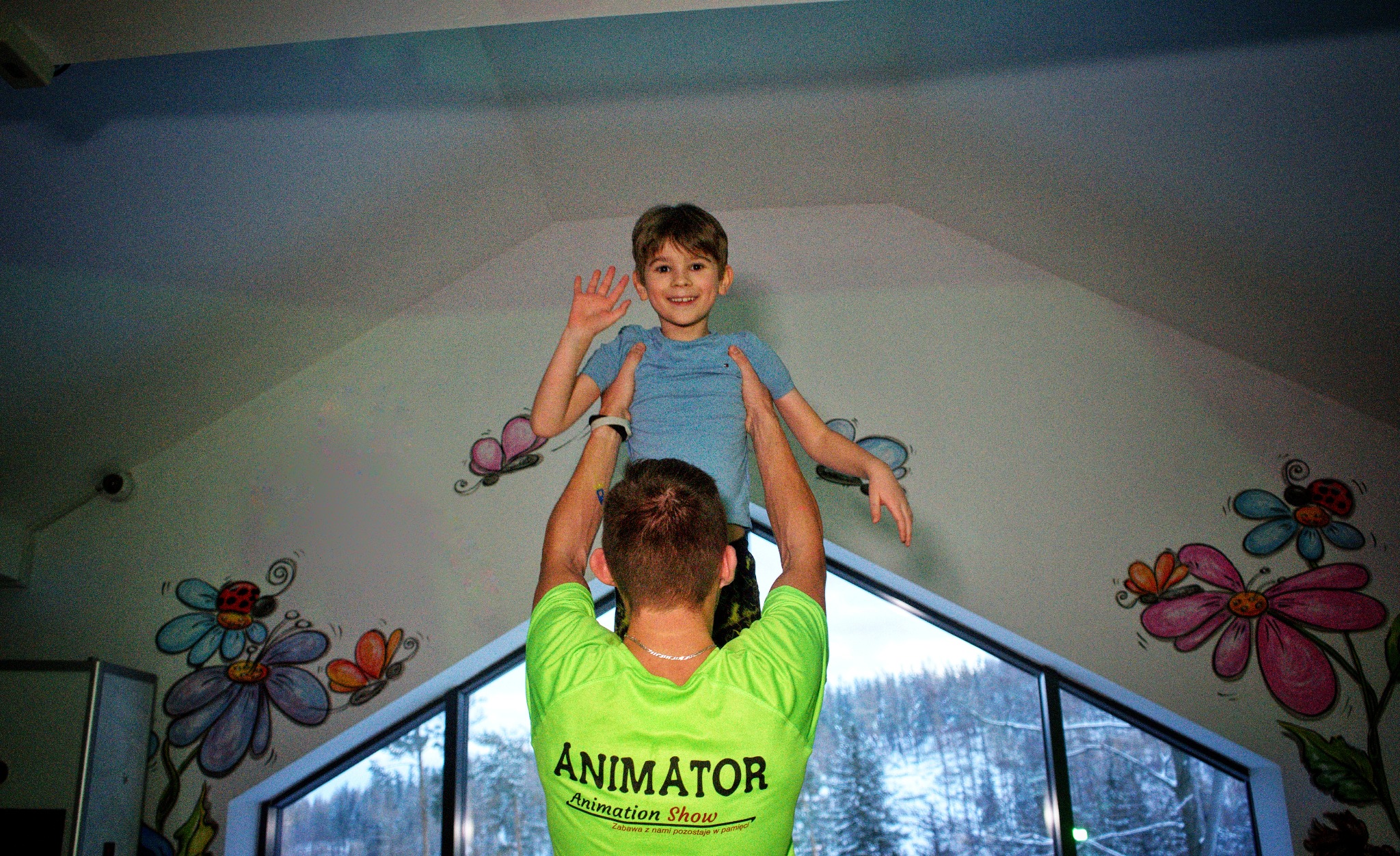 Mężczyzna w neonowej koszulce z napisem 'Animator Animation Show' podnosi uśmiechniętego chłopca na tle ściany z rysunkami kwiatów i motyli oraz okna z widokiem na zimowy krajobraz.