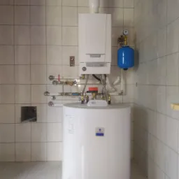 Biały piec gazowy kondensacyjny z podgrzewaczem wody, niebieskim naczyniem przeponowym i rurami w pomieszczeniu wyłożonym kwadratowymi, beżowymi płytkami ceramicznymi.