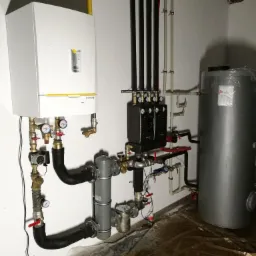 Biały piec grzewczy De Dietrich z żółtymi akcentami, czarne i szare rury instalacji grzewczej, szary zasobnik na wodę, widoczne manometry i zawory, folia ochronna na podłodze.