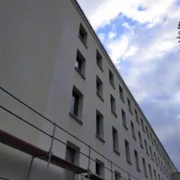 Długi budynek w trakcie prac elewacyjnych, widoczne rusztowania i fragmenty pomalowanej ściany, tło z niebem i chmurami.