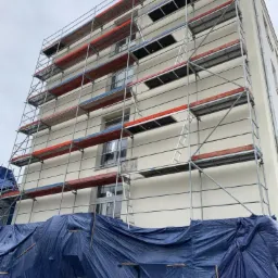 Budynek w trakcie ocieplania elewacji, widoczne rusztowania i plandeka zabezpieczająca teren prac.