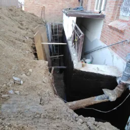 Wykop fundamentowy z zabezpieczoną ścianą, widoczna drabina zejściowa i fragment instalacji rurowej na tle ceglanego budynku.