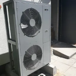 Szara, dwuwentylatorowa pompa ciepła zamontowana na betonowej podstawie przed budynkiem z widocznymi rurami doprowadzającymi i otwartymi drzwiami wejściowymi.