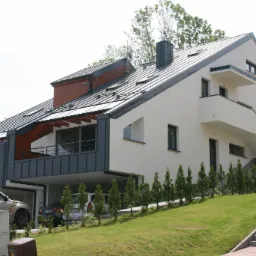 Nowoczesny dom dwurodzinny z płaskim dachem, wykończony w bieli i szarości, z balkonami i podcieniami garażowymi, otoczony trawnikiem i młodymi tujami.