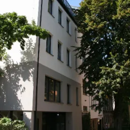 Nowoczesny budynek usługowy z białymi ścianami, czarnymi ramami okiennymi i balkonem z czarną balustradą, częściowo przysłonięty drzewami.